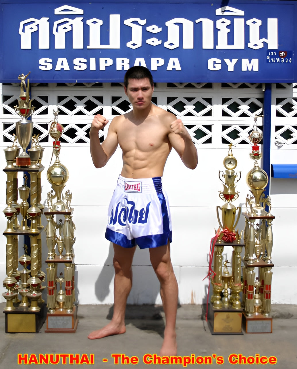 Thai boxer proudly displays his trophies in front of his traditional gym, wearing high-quality satin Hanuthai Blue and White Classic Muay Thai Boxing Shorts.