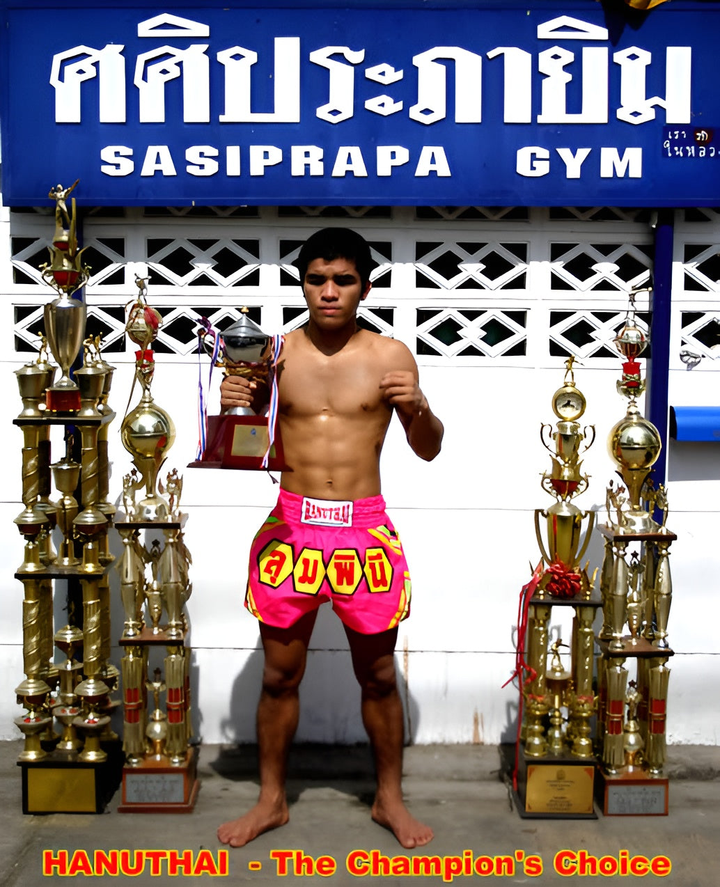 A man holding trophies in front of a gym, proudly displaying his achievements in fighting while wearing Hanuthai's Lumpini Pink Muay Thai Boxing Shorts.